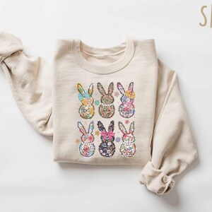 May include: A beige sweatshirt with a floral print of six bunnies. The bunnies are arranged in two rows of three. The sweatshirt has a crew neck and long sleeves.
