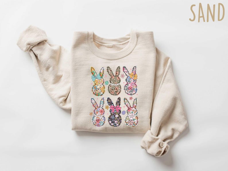 May include: A beige sweatshirt with a floral print of six bunnies. The bunnies are arranged in two rows of three. The sweatshirt has a crew neck and long sleeves.