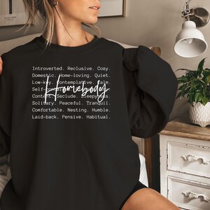 May include: Black sweatshirt with white text that reads "Introverted. Reclusive. Cozy. Domestic. Home-loving. Quiet. Low-key. Contemplative. Calm. Self-Homebody Content. Seclude. Sleepy. Solitary. Peaceful. Tranquil. Comfortable. Nesting. Humble. Laid-back. Pensive. Habitual."