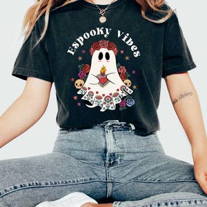 May include: A black t-shirt with a white ghost wearing a floral crown and holding a heart with a flame. The text "Spooky Vibes" is written in a retro font around the ghost.