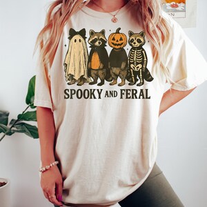 May include: Cream-colored t-shirt featuring a Halloween-themed graphic. The design includes a ghost, two raccoons, and a pumpkin-headed figure. The text "SPOOKY AND FERAL" is printed below the characters. The shirt is a casual, unisex style.