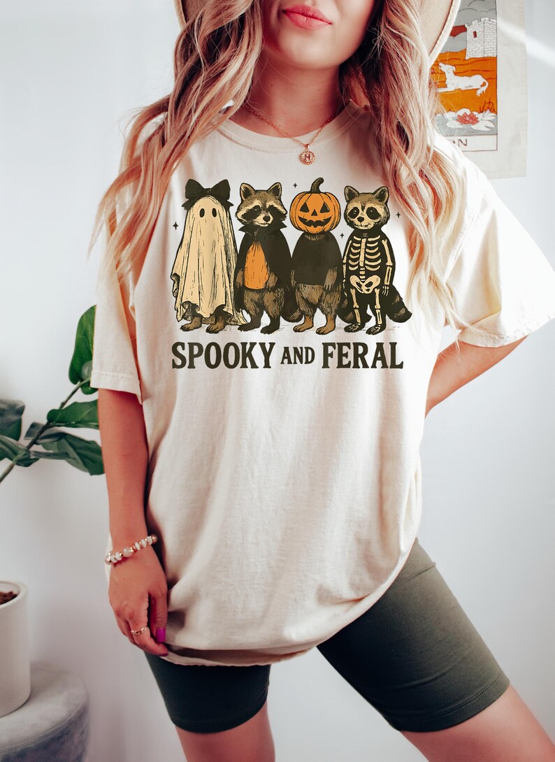 May include: Cream-colored t-shirt featuring a Halloween-themed graphic. The design includes a ghost, two raccoons, and a pumpkin-headed figure. The text "SPOOKY AND FERAL" is printed below the characters. The shirt is a casual, unisex style.