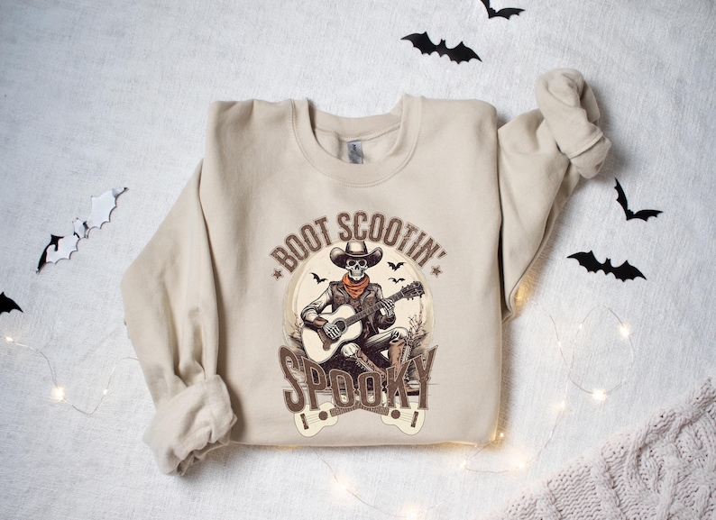 May include: A beige sweatshirt with a graphic print of a skeleton playing a guitar. The text on the sweatshirt reads "Boot Scootin' Spooky".