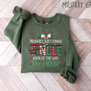 Peut inclure: Sweat-shirt vert militaire avec l'inscription "Probably Just Gonna Jingle Some Of The Way I'm Tired" dans un design festif. Le mot "Jingle" est d&eacute;cor&eacute; de guirlandes lumineuses, de carreaux et de rayures. Un n&oelig;ud et du houx ornent le haut.