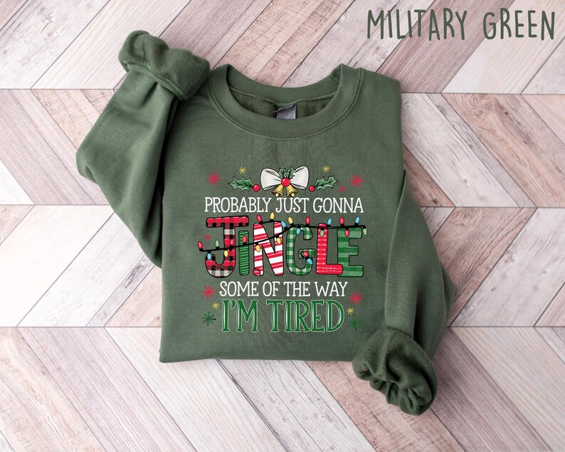 Peut inclure: Sweat-shirt vert militaire avec l'inscription "Probably Just Gonna Jingle Some Of The Way I'm Tired" dans un design festif. Le mot "Jingle" est d&eacute;cor&eacute; de guirlandes lumineuses, de carreaux et de rayures. Un n&oelig;ud et du houx ornent le haut.