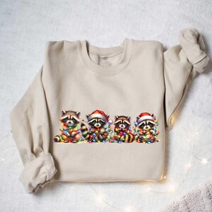 May include: A beige sweatshirt with a graphic of four raccoons wearing Santa hats and holding Christmas lights. The raccoons are surrounded by colorful Christmas lights.