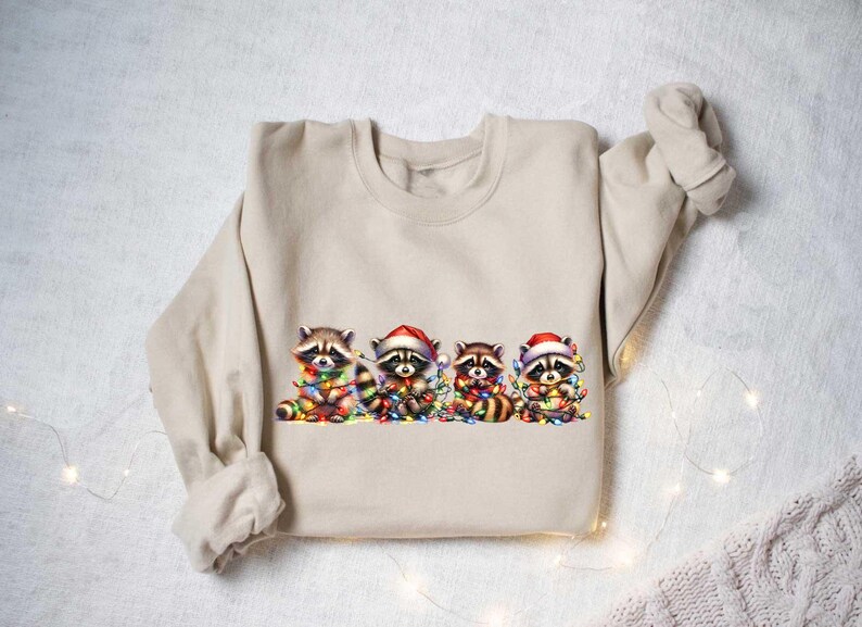 May include: A beige sweatshirt with a graphic of four raccoons wearing Santa hats and holding Christmas lights. The raccoons are surrounded by colorful Christmas lights.