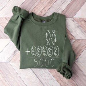 May include: A green sweatshirt with a white graphic of two fish and five loaves of bread. The text "5000" is below the graphic.