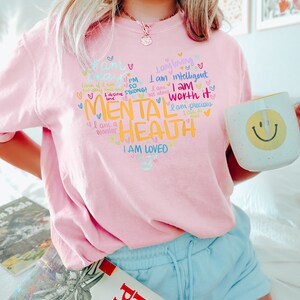 Op de afbeelding: Roze T-shirt met een grafisch ontwerp met de tekst "Ik ben sterk! Ik ben het waard. Geestelijke gezondheid. Ik ben geliefd." Het grafische ontwerp is in verschillende kleuren en bevat harten en andere krabbels.