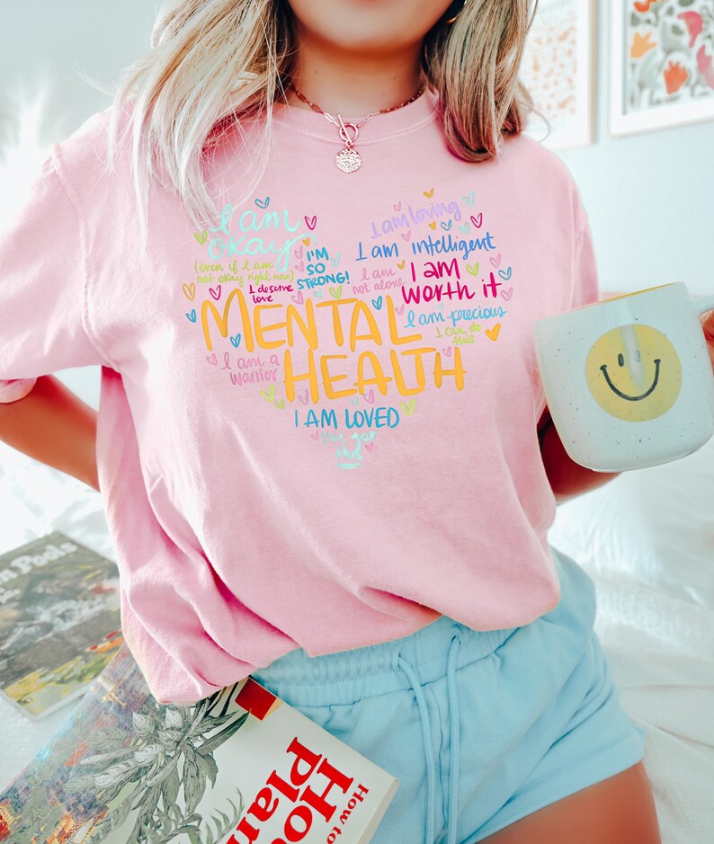 Op de afbeelding: Roze T-shirt met een grafisch ontwerp met de tekst "Ik ben sterk! Ik ben het waard. Geestelijke gezondheid. Ik ben geliefd." Het grafische ontwerp is in verschillende kleuren en bevat harten en andere krabbels.