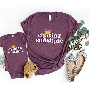 May include: A set of two purple t-shirts with a sun and text. The larger shirt says "chasing sunshine" and the smaller shirt says "mama's sunshine".