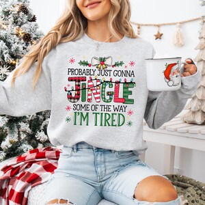 Peut inclure: Un sweat-shirt gris clair avec le texte "Probably Just Gonna Jingle Some of the Way I'm Tired" dans une police festive. Le mot "Jingle" est en carreaux et vert. Une tasse blanche avec un motif de P&egrave;re No&euml;l est tenue &agrave; la main.