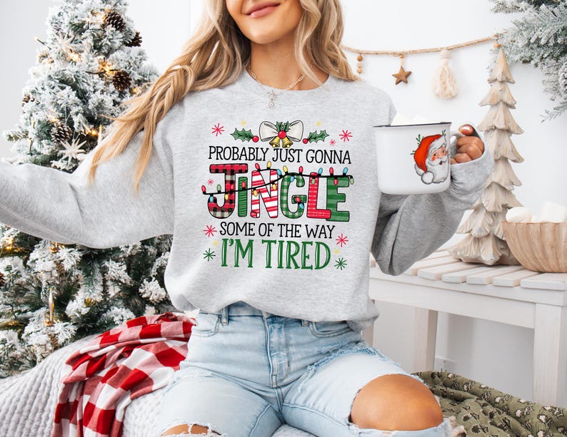 Peut inclure: Un sweat-shirt gris clair avec le texte "Probably Just Gonna Jingle Some of the Way I'm Tired" dans une police festive. Le mot "Jingle" est en carreaux et vert. Une tasse blanche avec un motif de P&egrave;re No&euml;l est tenue &agrave; la main.