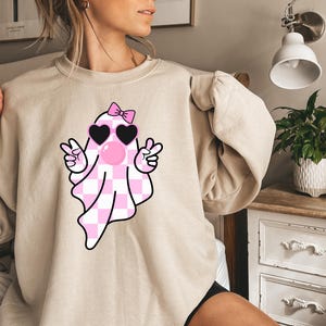 K&ouml;nnte beinhalten: Ein hellbeiger Pullover mit einem rosa-wei&szlig; karierten Geist, der eine rosa Sonnenbrille und eine rosa Schleife tr&auml;gt. Der Geist zeigt mit beiden H&auml;nden ein Peace-Zeichen.