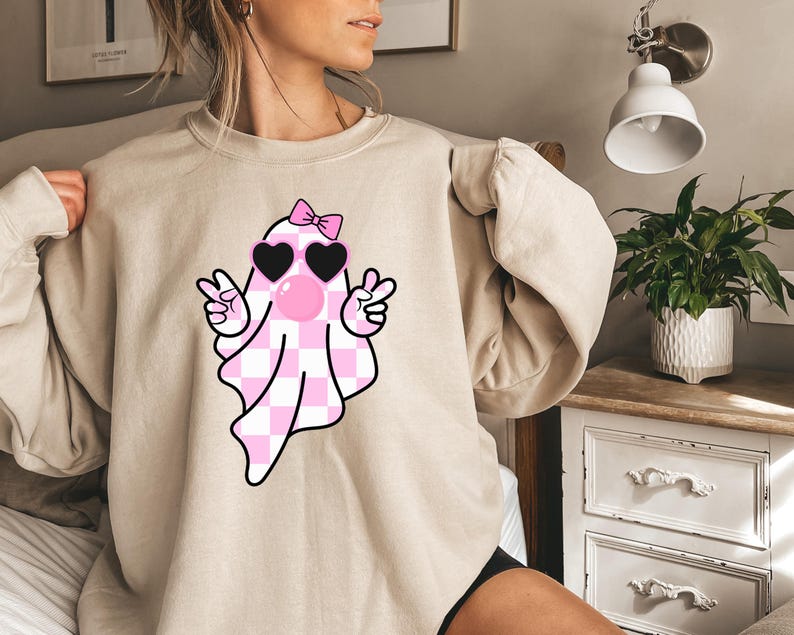 K&ouml;nnte beinhalten: Ein hellbeiger Pullover mit einem rosa-wei&szlig; karierten Geist, der eine rosa Sonnenbrille und eine rosa Schleife tr&auml;gt. Der Geist zeigt mit beiden H&auml;nden ein Peace-Zeichen.