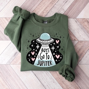 以下が含まれることがあります： 「Boys Go To Jupiter」というテキストとピンクのハートが描かれたUFOの白黒のグラフィックが施された緑色のスウェットシャツです。