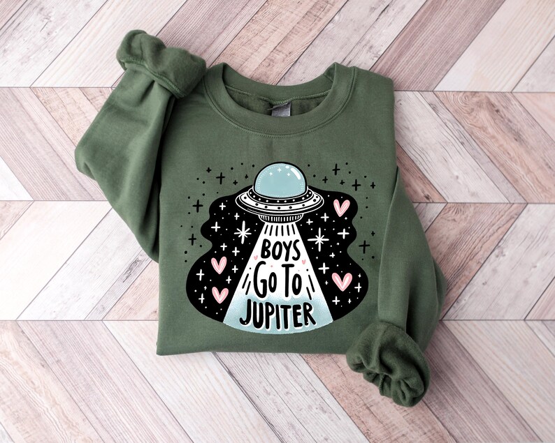 以下が含まれることがあります： 「Boys Go To Jupiter」というテキストとピンクのハートが描かれたUFOの白黒のグラフィックが施された緑色のスウェットシャツです。