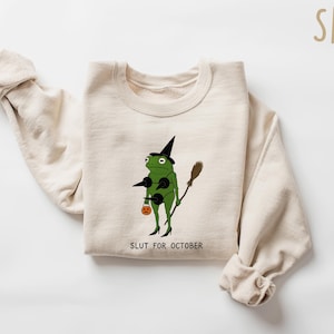 May include: Sand-colored sweatshirt featuring a green frog dressed as a witch, holding a broom and a pumpkin-shaped treat bag. The text "SLUT FOR OCTOBER" is printed below the frog. The sweatshirt has long sleeves.