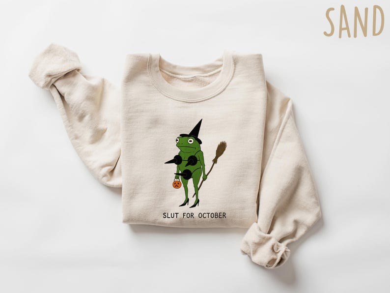 May include: Sand-colored sweatshirt featuring a green frog dressed as a witch, holding a broom and a pumpkin-shaped treat bag. The text "SLUT FOR OCTOBER" is printed below the frog. The sweatshirt has long sleeves.
