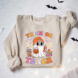 May include: A beige sweatshirt with a graphic of a ghost holding a pumpkin and a pencil. The text "Trick or Teach" is printed in colorful letters.
