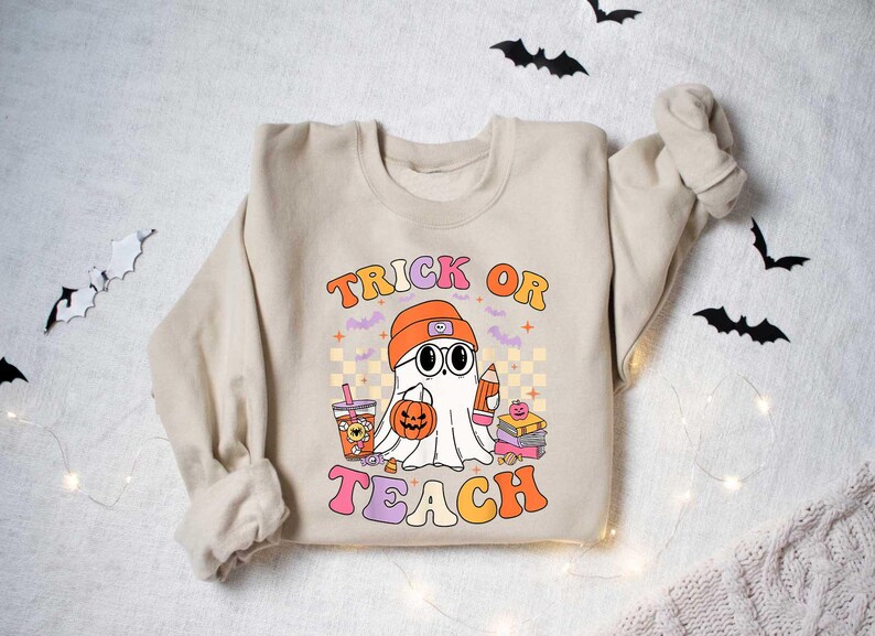 May include: A beige sweatshirt with a graphic of a ghost holding a pumpkin and a pencil. The text "Trick or Teach" is printed in colorful letters.