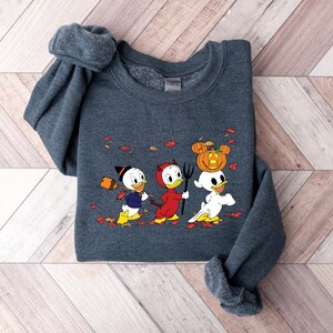 May include: A dark grey sweatshirt with a Halloween-themed design featuring three cartoon ducks. One duck is dressed as a devil, another is wearing a witch's hat, and the third is carrying a pumpkin on its head. The ducks are walking on a path with autumn leaves.