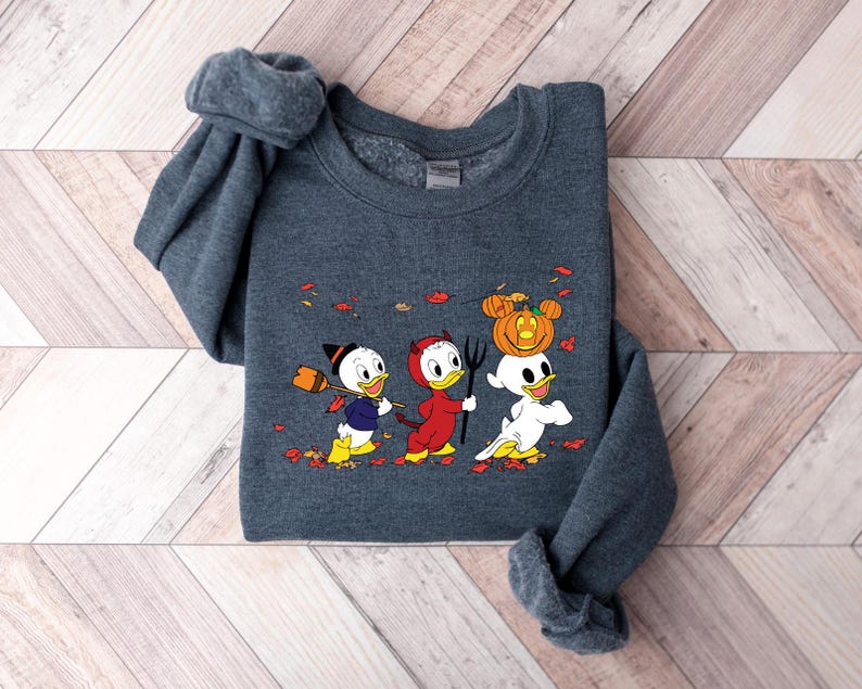 May include: A dark grey sweatshirt with a Halloween-themed design featuring three cartoon ducks. One duck is dressed as a devil, another is wearing a witch's hat, and the third is carrying a pumpkin on its head. The ducks are walking on a path with autumn leaves.