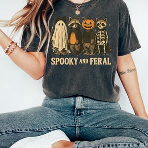 May include: Dark gray t-shirt with a Halloween-themed graphic. The design features a ghost, raccoon, pumpkin, and skeleton raccoon, with the words "Spooky and Feral" in a vintage font. The shirt is worn with blue jeans and white sneakers.