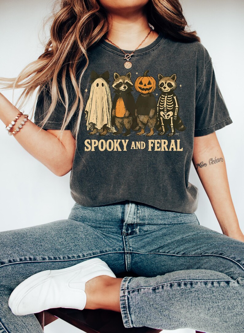 May include: Dark gray t-shirt with a Halloween-themed graphic. The design features a ghost, raccoon, pumpkin, and skeleton raccoon, with the words "Spooky and Feral" in a vintage font. The shirt is worn with blue jeans and white sneakers.