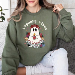 May include: A green sweatshirt with a white ghost wearing a floral crown and holding a heart. The text "Spooky Vibes" is printed on the sweatshirt.