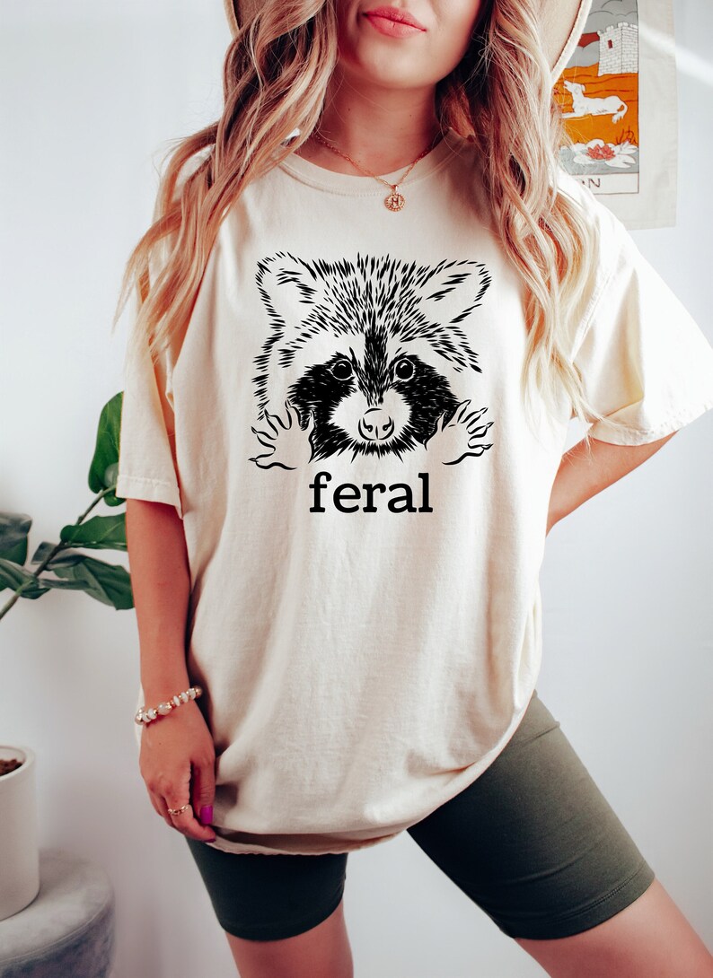 以下が含まれることがあります： クリーム色のTシャツに、黒と白のラクーンドッグの顔のイラストが描かれています。ラクーンドッグの顔はTシャツの後ろから顔を出しています。Tシャツの下には「feral」という文字が印刷されています。