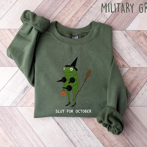May include: Military green sweatshirt with a graphic of a frog wearing a witch hat, holding a broom and a jack-o'-lantern. The text "SLUT FOR OCTOBER" is printed below the frog. The sweatshirt is folded on a light wood-grain surface.