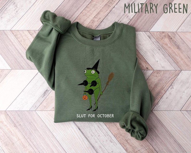 May include: Military green sweatshirt with a graphic of a frog wearing a witch hat, holding a broom and a jack-o'-lantern. The text "SLUT FOR OCTOBER" is printed below the frog. The sweatshirt is folded on a light wood-grain surface.