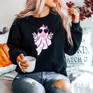 K&ouml;nnte beinhalten: Schwarzer Pullover mit einem rosa-wei&szlig; karierten Geist, der eine rosa Herz-Sonnenbrille und eine rosa Schleife tr&auml;gt. Der Geist macht mit beiden H&auml;nden ein Peace-Zeichen.