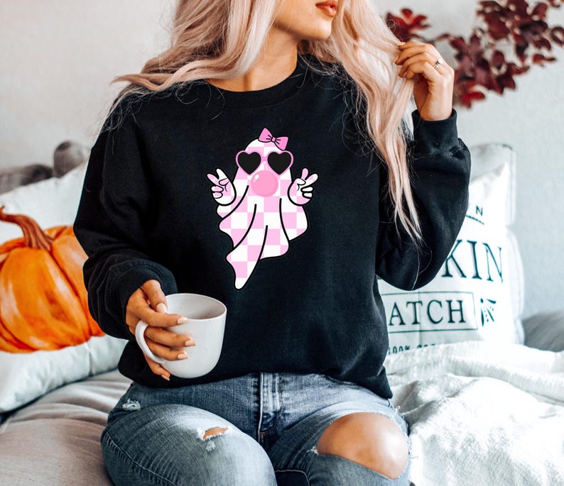 K&ouml;nnte beinhalten: Schwarzer Pullover mit einem rosa-wei&szlig; karierten Geist, der eine rosa Herz-Sonnenbrille und eine rosa Schleife tr&auml;gt. Der Geist macht mit beiden H&auml;nden ein Peace-Zeichen.
