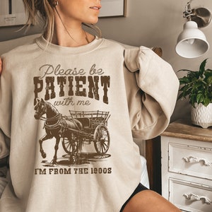 K&ouml;nnte beinhalten: Ein beiger Pullover mit einem braunen Grafikdruck eines Pferdes, das einen Wagen zieht. Der Text auf dem Pullover lautet "Please be PATIENT with me I'M FROM THE 1900s".