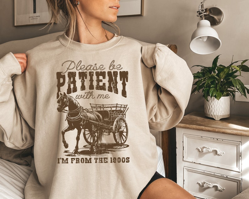 K&ouml;nnte beinhalten: Ein beiger Pullover mit einem braunen Grafikdruck eines Pferdes, das einen Wagen zieht. Der Text auf dem Pullover lautet "Please be PATIENT with me I'M FROM THE 1900s".