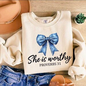 May include: Cream-colored sweatshirt with a blue bow graphic and the text "She is worthy PROVERBS 31". The sweatshirt is laid flat with blue jeans, a brown hat, and sandals.