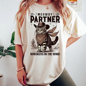 K&ouml;nnte beinhalten: Ein beiges T-Shirt mit einem Aufdruck eines Katers mit Cowboyhut und Bandana. Der Text "MEOWDY PARTNER" und "WHISKERS IN THE WIND" ist auf dem Shirt gedruckt.