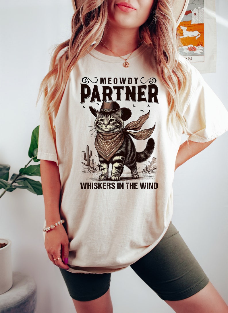 K&ouml;nnte beinhalten: Ein beiges T-Shirt mit einem Aufdruck eines Katers mit Cowboyhut und Bandana. Der Text "MEOWDY PARTNER" und "WHISKERS IN THE WIND" ist auf dem Shirt gedruckt.