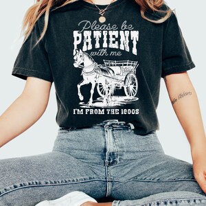 K&ouml;nnte beinhalten: Ein schwarzes T-Shirt mit einer wei&szlig;en Grafik einer Pferdekutsche und dem Text "Please be PATIENT with me I'M FROM THE 1900s".