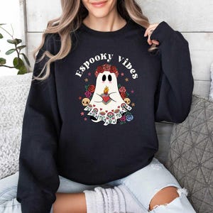 May include: Black sweatshirt with a white ghost wearing a floral crown and holding a heart. The text "Spooky Vibes" is printed in white above the ghost.