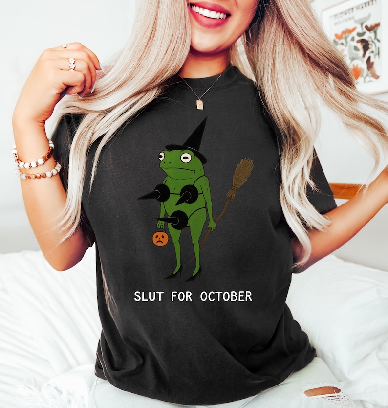 May include: Black t-shirt with a green frog wearing a witch hat, a black bikini, and holding a broom and a jack-o'-lantern. The text "SLUT FOR OCTOBER" is printed in white on the shirt. The frog is the central graphic.