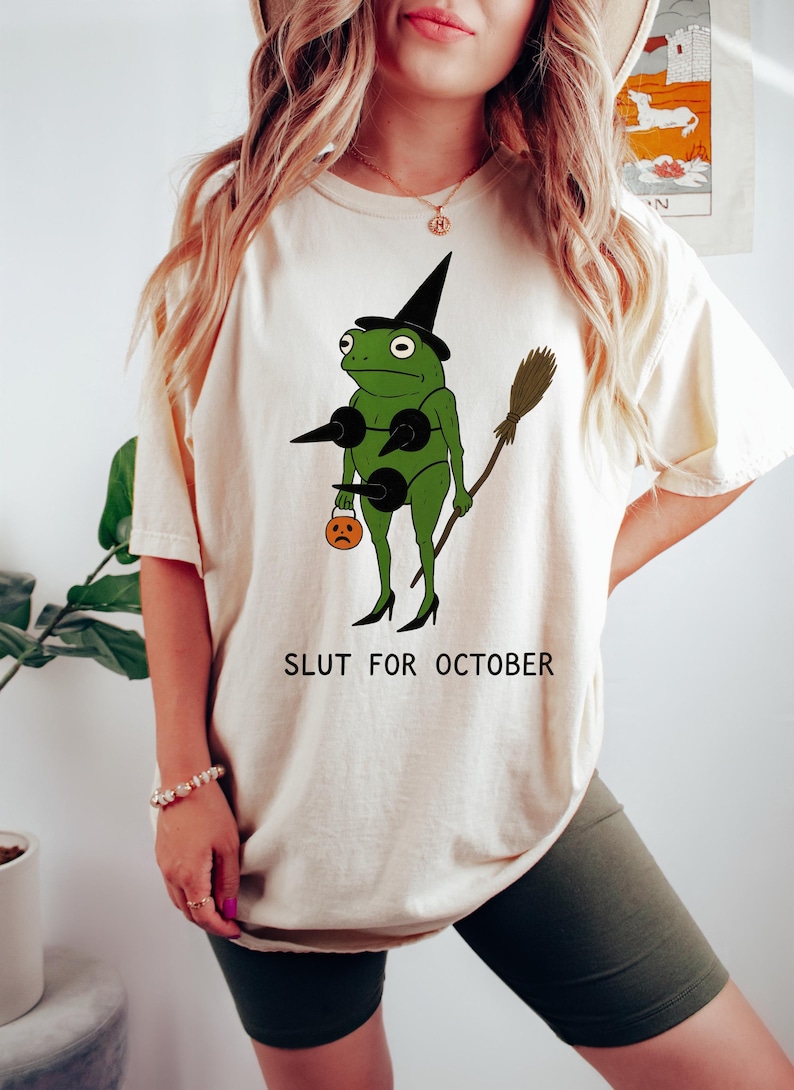 May include: Cream-colored t-shirt with a green frog graphic in a witch costume, holding a broom and a jack-o'-lantern. The text "SLUT FOR OCTOBER" is printed below the graphic. The frog wears a black witch hat and high heels.