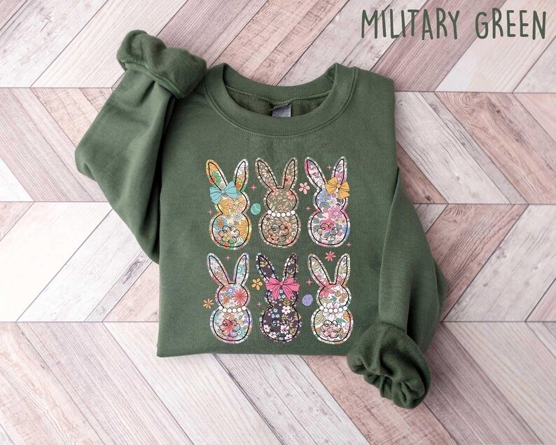 May include: A green sweatshirt with a floral print of six bunnies. The bunnies are wearing bows and have a vintage floral pattern. The sweatshirt has the text "MILITARY GREEN" printed on the top right corner.