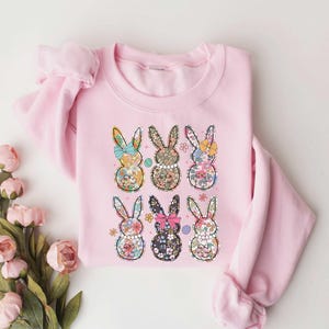 May include: Pink sweatshirt with a floral pattern of six bunnies. Each bunny has a bow and is surrounded by flowers.