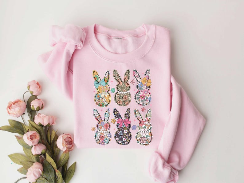 May include: Pink sweatshirt with a floral pattern of six bunnies. Each bunny has a bow and is surrounded by flowers.