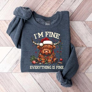 May include: A dark blue sweatshirt with a Christmas-themed design featuring a Highland cow wearing a Santa hat and surrounded by Christmas lights. The text "I'm Fine Everything is Fine" is printed on the sweatshirt.