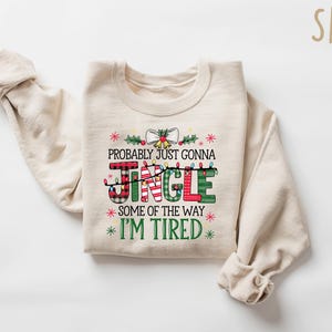 Peut inclure: Un sweat-shirt de couleur sable avec l'inscription "Probably Just Gonna Jingle Some of the Way I'm Tired" dans un design festif. Le mot "JINGLE" est en rouge, vert et motifs &eacute;cossais, avec des guirlandes lumineuses. Un sweat-shirt sur le th&egrave;me des f&ecirc;tes.
