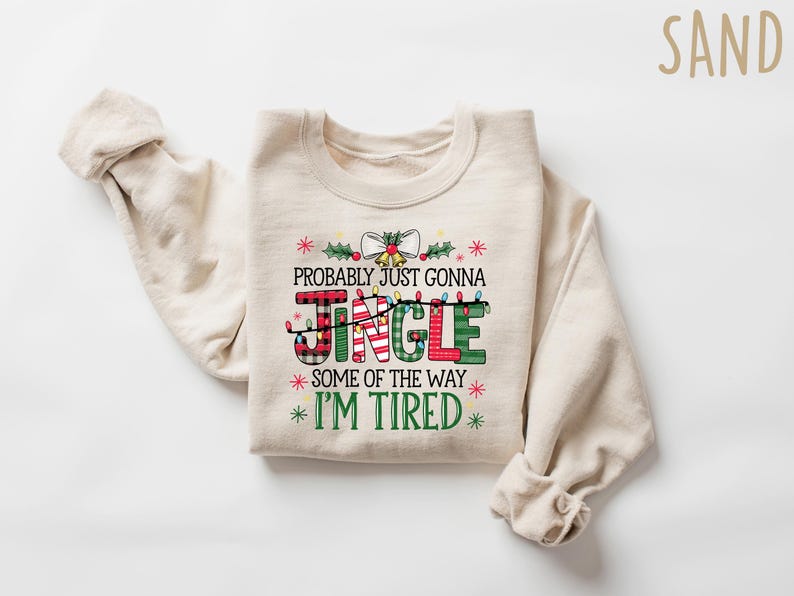 Peut inclure: Un sweat-shirt de couleur sable avec l'inscription "Probably Just Gonna Jingle Some of the Way I'm Tired" dans un design festif. Le mot "JINGLE" est en rouge, vert et motifs &eacute;cossais, avec des guirlandes lumineuses. Un sweat-shirt sur le th&egrave;me des f&ecirc;tes.